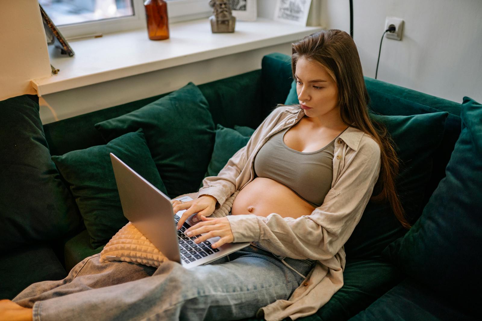 Expectant mother working on her laptop comfortably on a sofa, symbolizing remote work and digital nomad lifestyle.